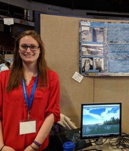 Image of Emily Blackwood standing in front of her Presentation at the UMaine Student Symposium. She is wearing a red long sleeve shirt.