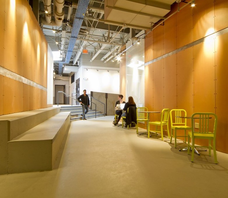 IMRC Center hallway with two students conversing at a small silver table. A third student walks toward them from a small flight of stars positioned in the corner of the hallway.