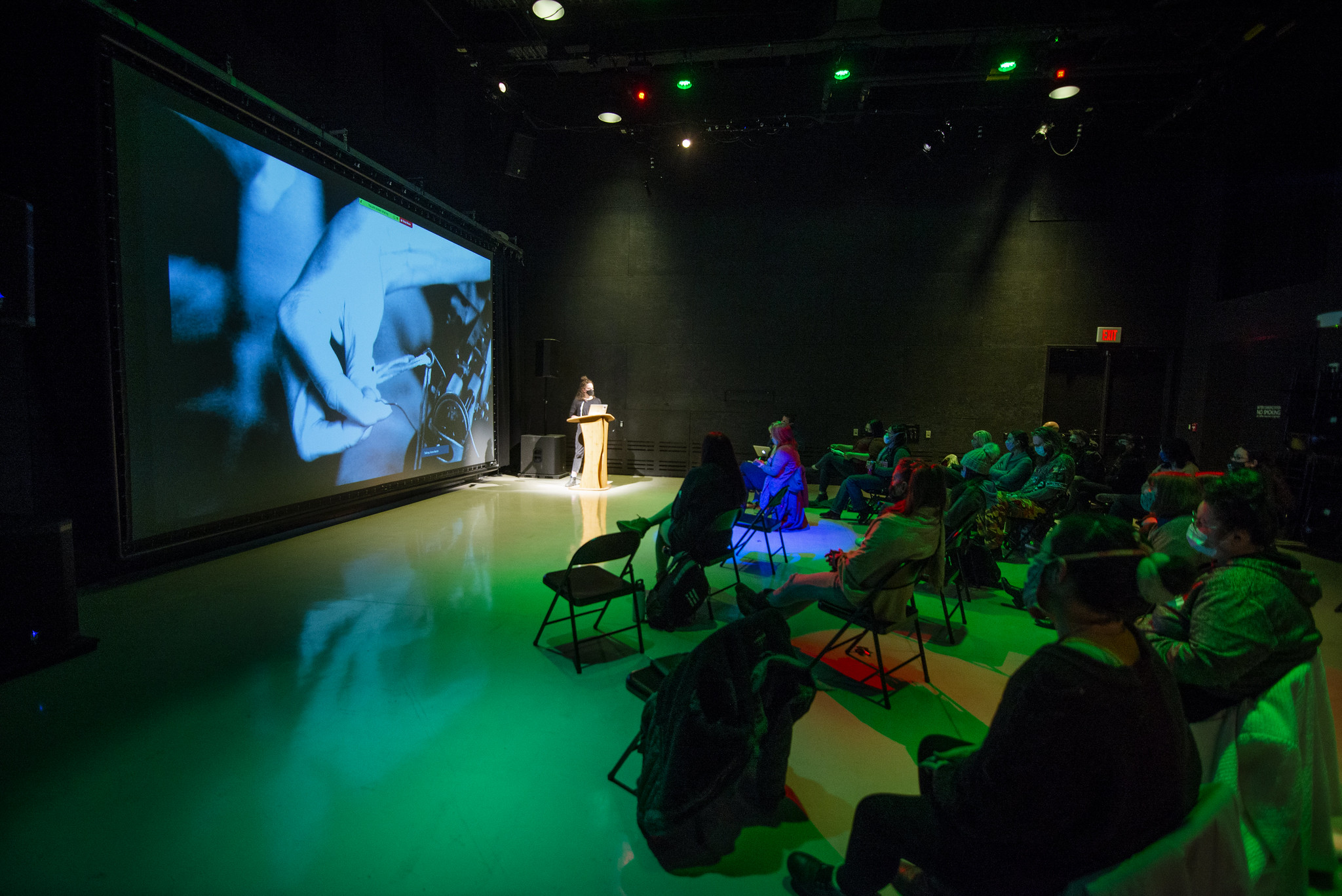 Kate Fogler presents inside the Adaptive Presentation and Performance Envrionment at IMRC Center. There is a large projection screen with an image of a hand pulling a needle through fabric. Green lighting from the ceiling of the room and students in chairs.