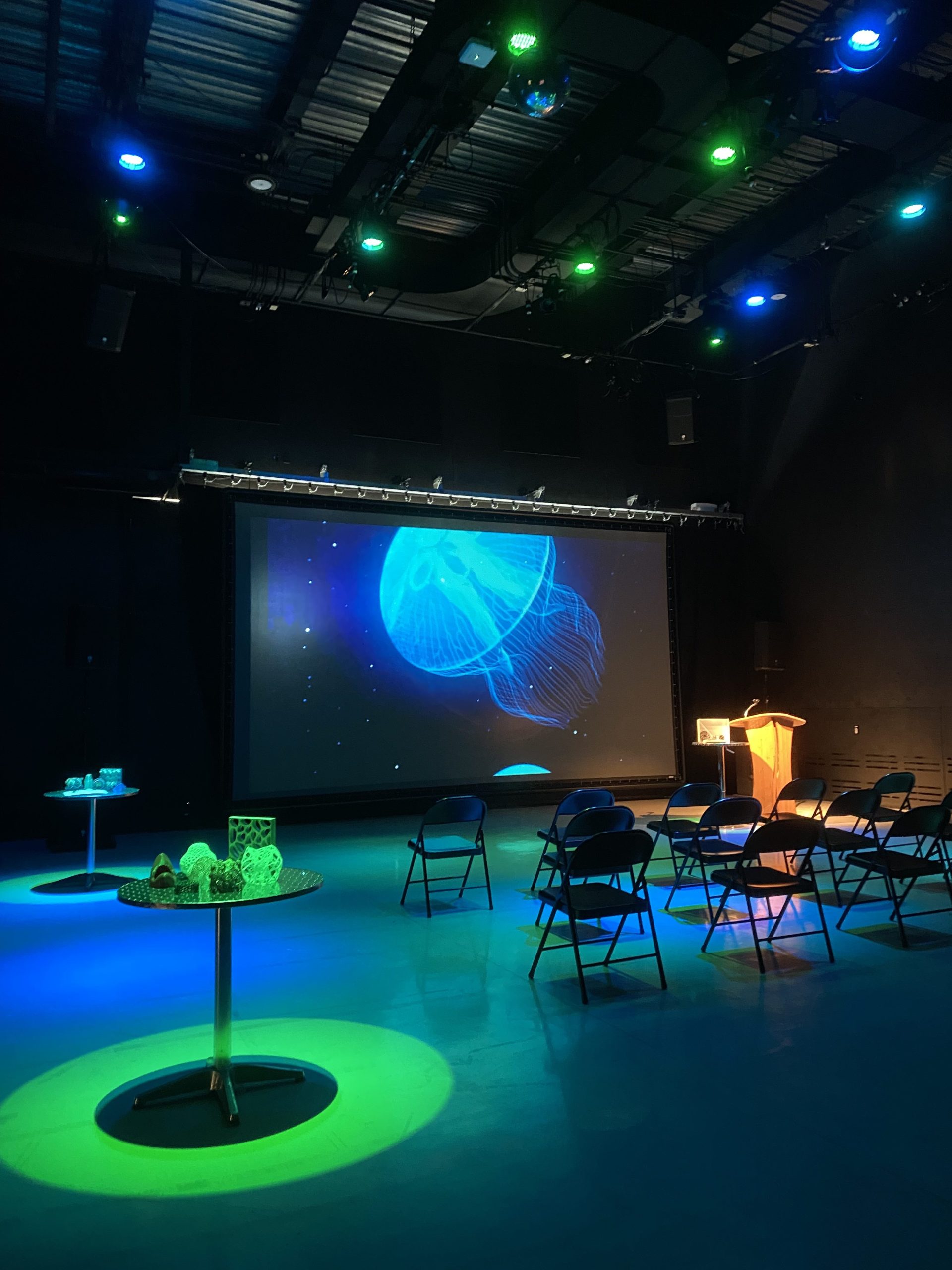 Green and blue theater lights shine on 3D printed objects in the IMRC Center's Adaptive Presentation and Performance Environment. A jellyfish swims on the projection screen.