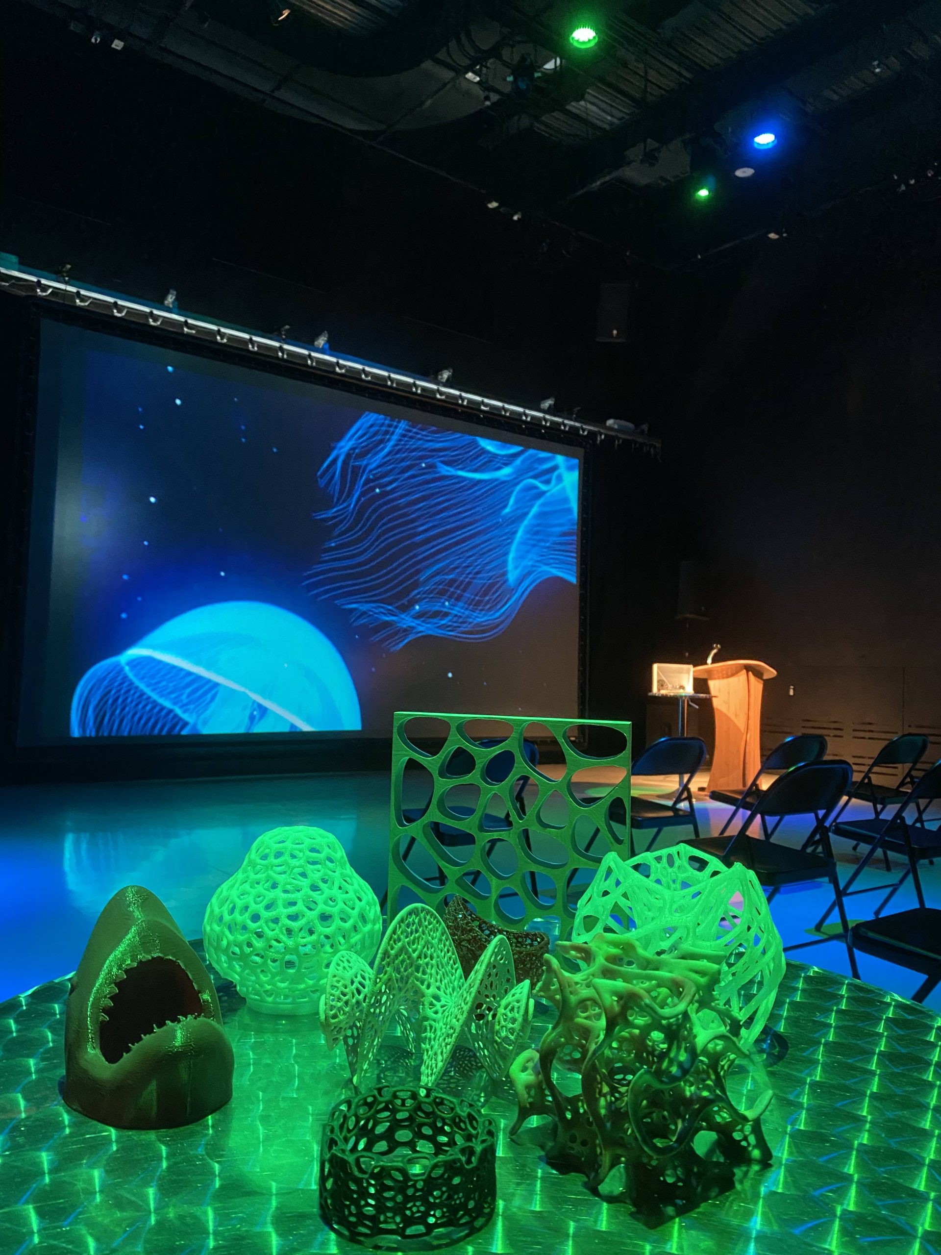 Green and blue theater lights shine on 3D printed objects in the IMRC Center's Adaptive Presentation and Performance Environment. A jellyfish swims on the projection screen.