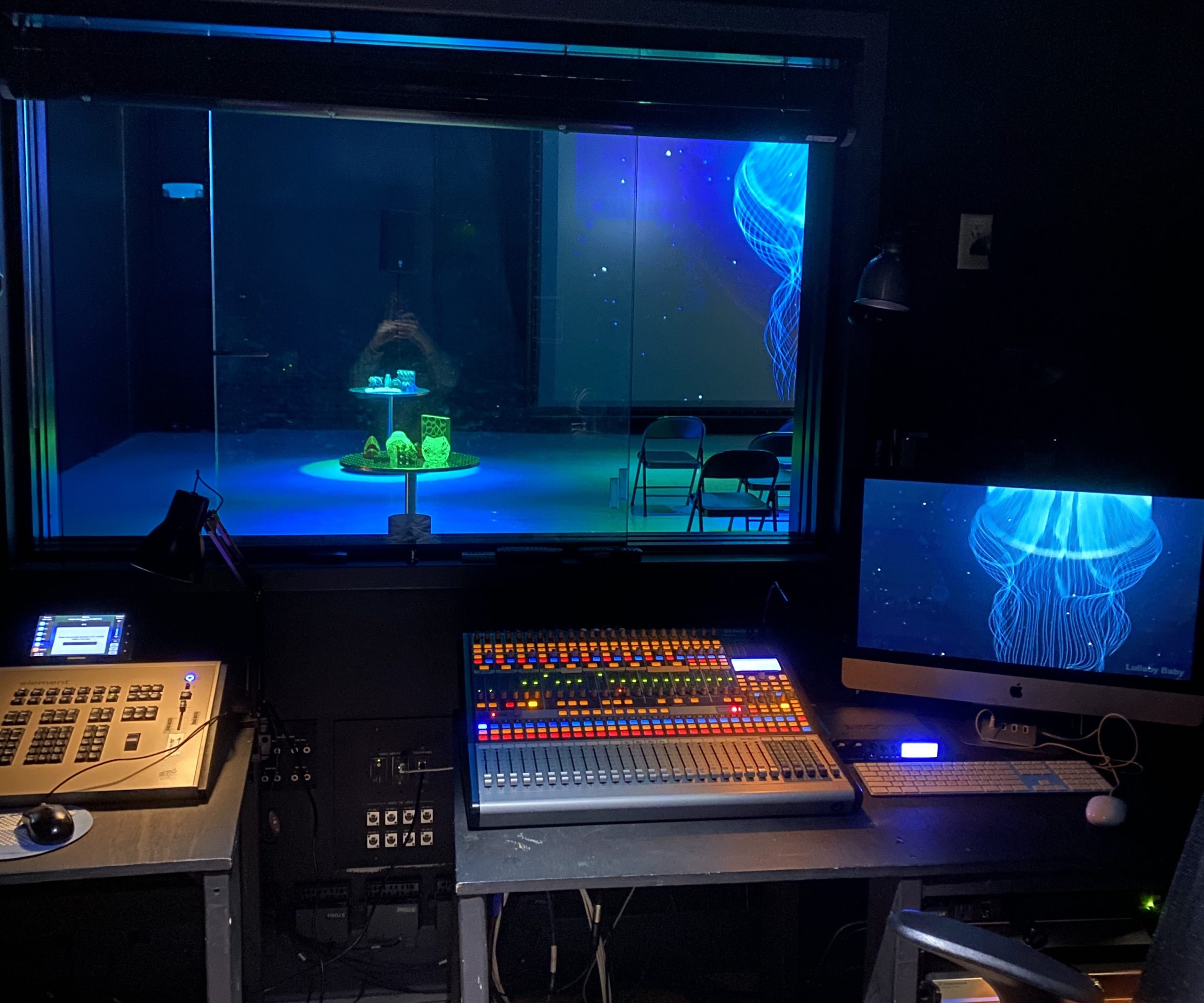 Green and blue theater lights shine on 3D printed objects in the IMRC Center's Adaptive Presentation and Performance Environment. A jellyfish swims on the projection screen. This is viewed from the perspective of looking through a window in the control booth.