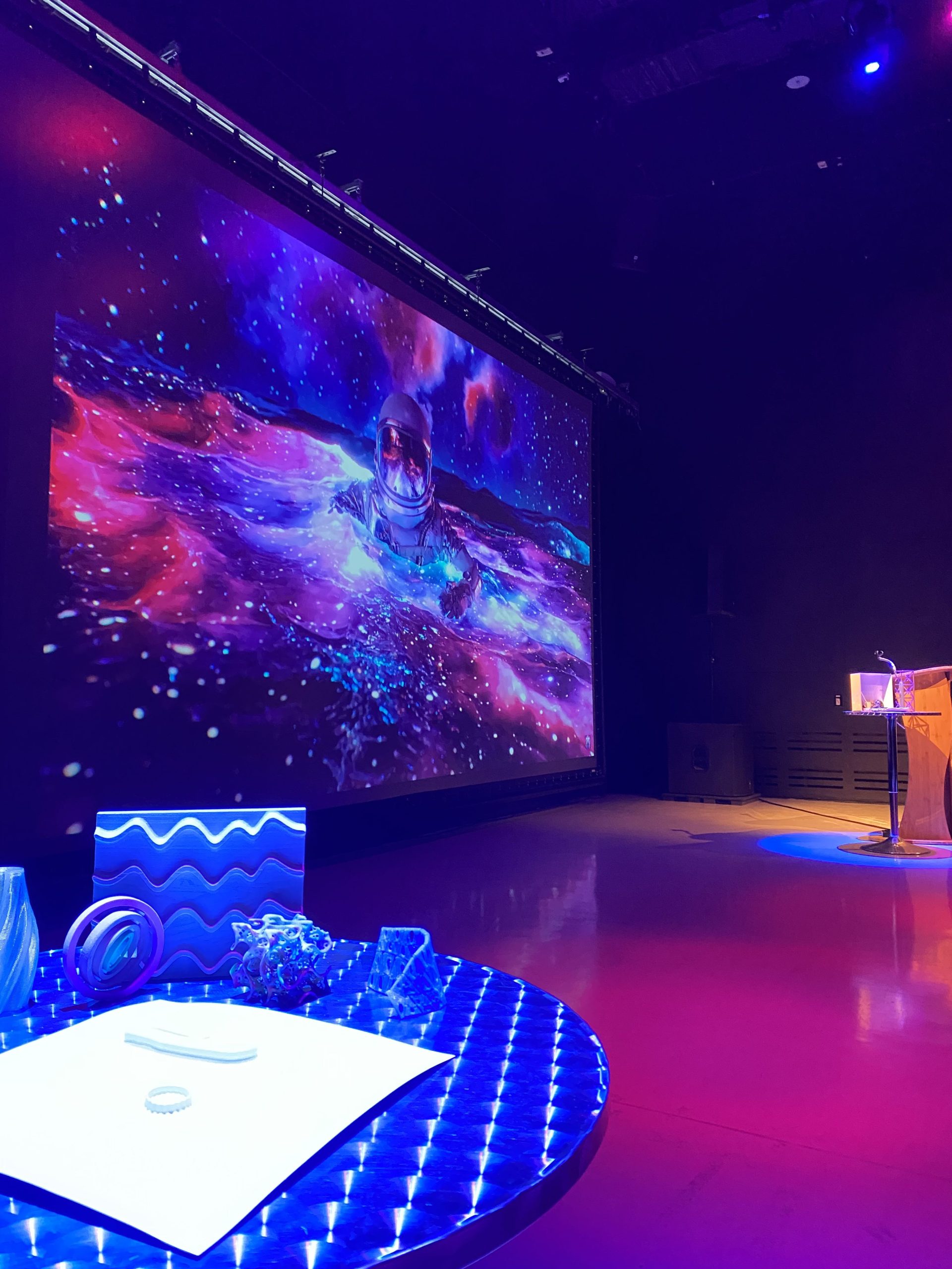 Purple and blue theater lights shine on 3D printed objects in the IMRC Center's Adaptive Presentation and Performance Environment. An astronaut floats on the projection screen.