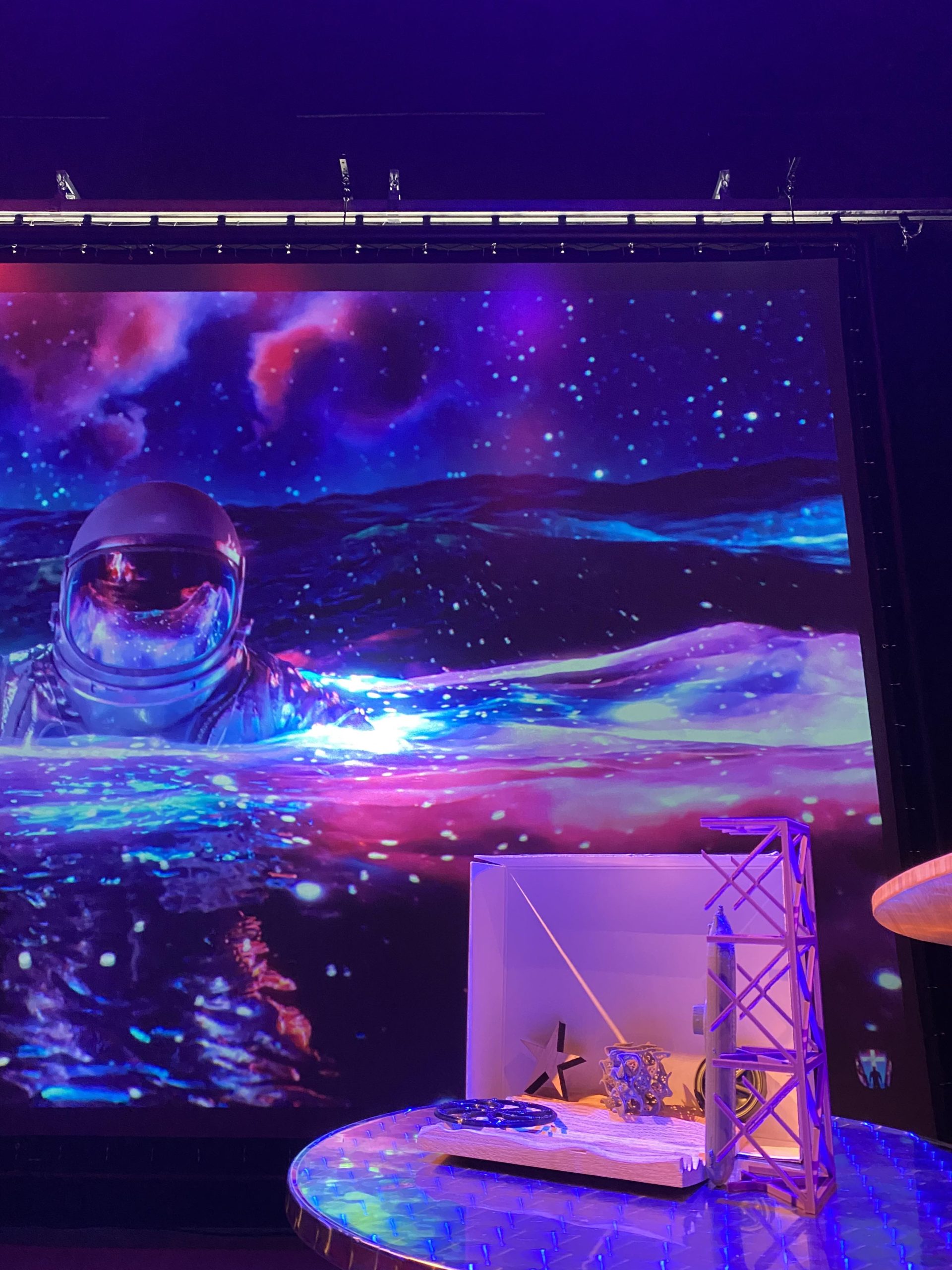 Purple and blue theater lights shine on 3D printed objects in the IMRC Center's Adaptive Presentation and Performance Environment. An astronaut floats on the projection screen.