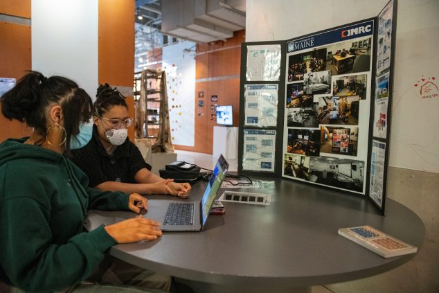 Two masked students work side by side at a round table in front of an IMRC Center folding banner