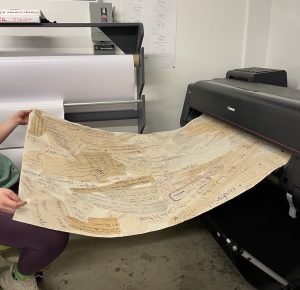 IMFA student Alex Rose holds a piece of fabric from off camera on the left as it prints through a Canon large format printer on the right. The piece is several feet long by several feet wide and shows a variety of handwritten notes and drawings