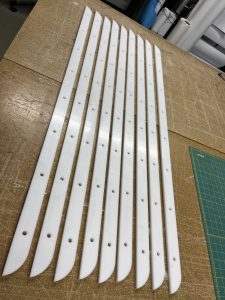 White acrylic jigs, several feet long by about an inch wide, are lined up next to each other on a workbench