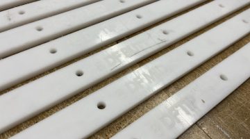 A close up shot of white acrylic jigs, several feet long by about an inch wide, are lined up next to each other on a workbench