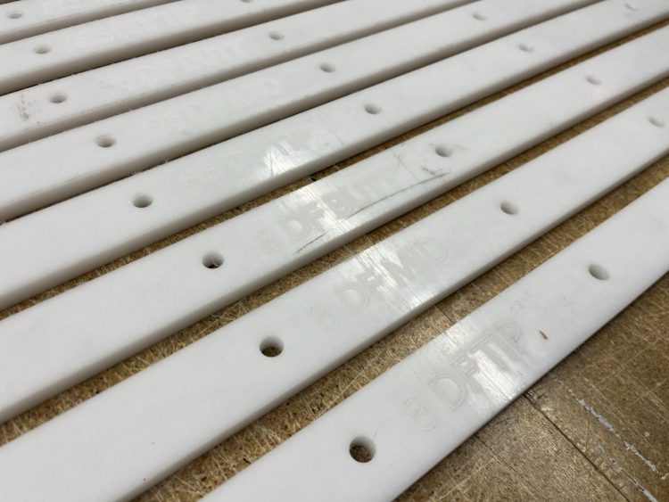 A close up shot of white acrylic jigs, several feet long by about an inch wide, are lined up next to each other on a workbench