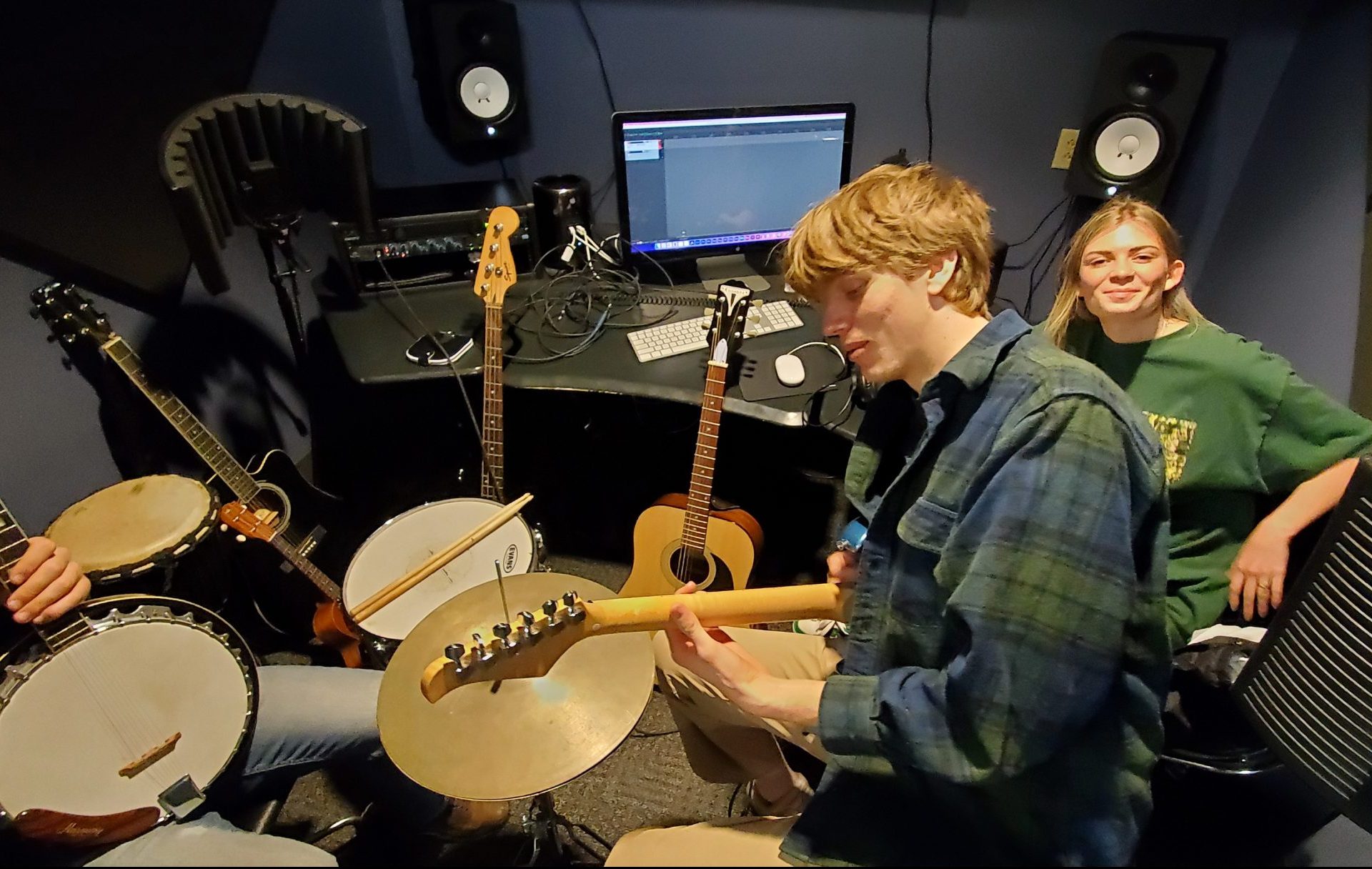 Phoenix is seated at the center of the image holding a guitar. At the left, a student holds a banjo, while another student behind Phoenix looks directly at the camera smiling. The students are seated in an audiovisual lab with a mixing program on a computer behind them.