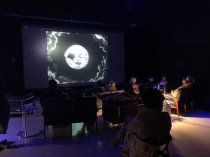 Guests at the IMRC Center are seated in the APPE Space, viewing an early film on the projection screen. The room is darkened, and a small lamp illuminates the right side of the image.