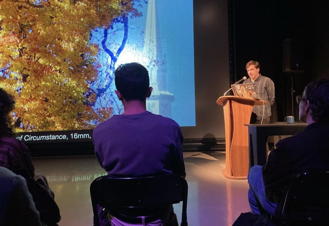 A speaker stands behind a wooden podium to the right of the image. Next to him is a large projection screen with an image of a tree with golden leaves, a blue sky, and a white steeple. People sit in shadow in folding chairs in the foreground.