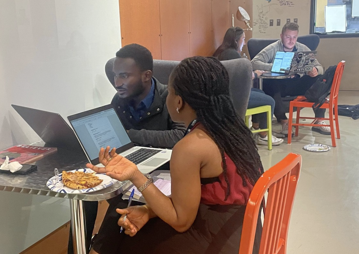 Four students are seated at two separate tables, looking at laptops with information that they are discussing.