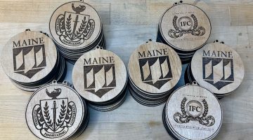 Eight stacks of laser-cut wooden medallions on a wooden work surface. The medallions have a variety of logos.