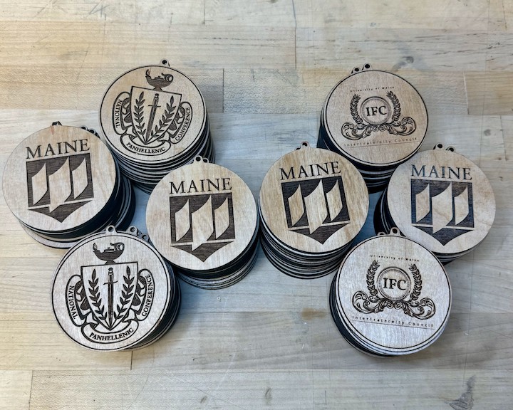 Eight stacks of laser-cut wooden medallions on a wooden work surface. The medallions have a variety of logos.