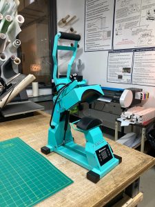 A bright teal tabletop hat press sits on top of a wooden work table.