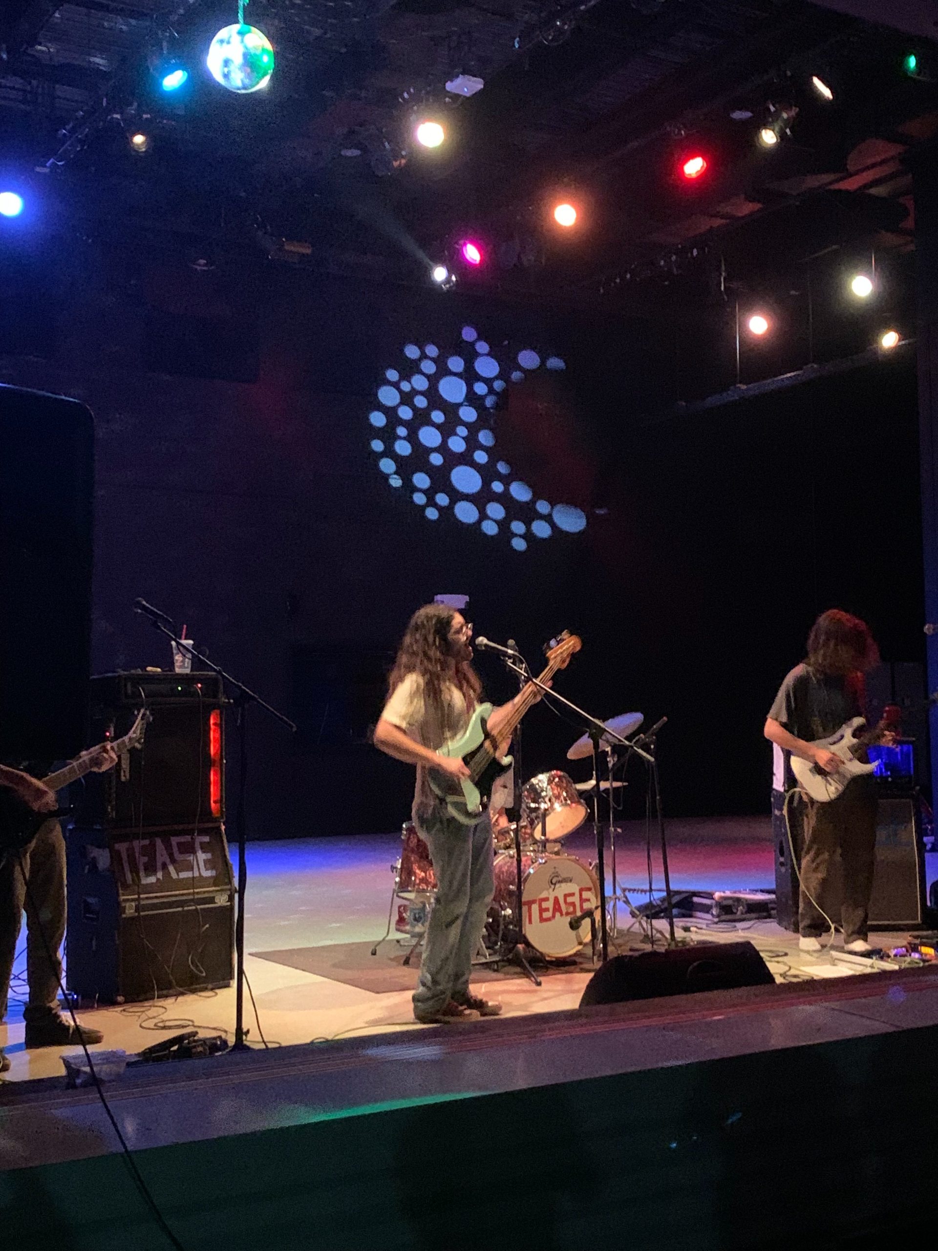 A band plays a live concert on a stage with theatrical lighting. Two of the musicians hold guitars. The central musician sings into a microphone.