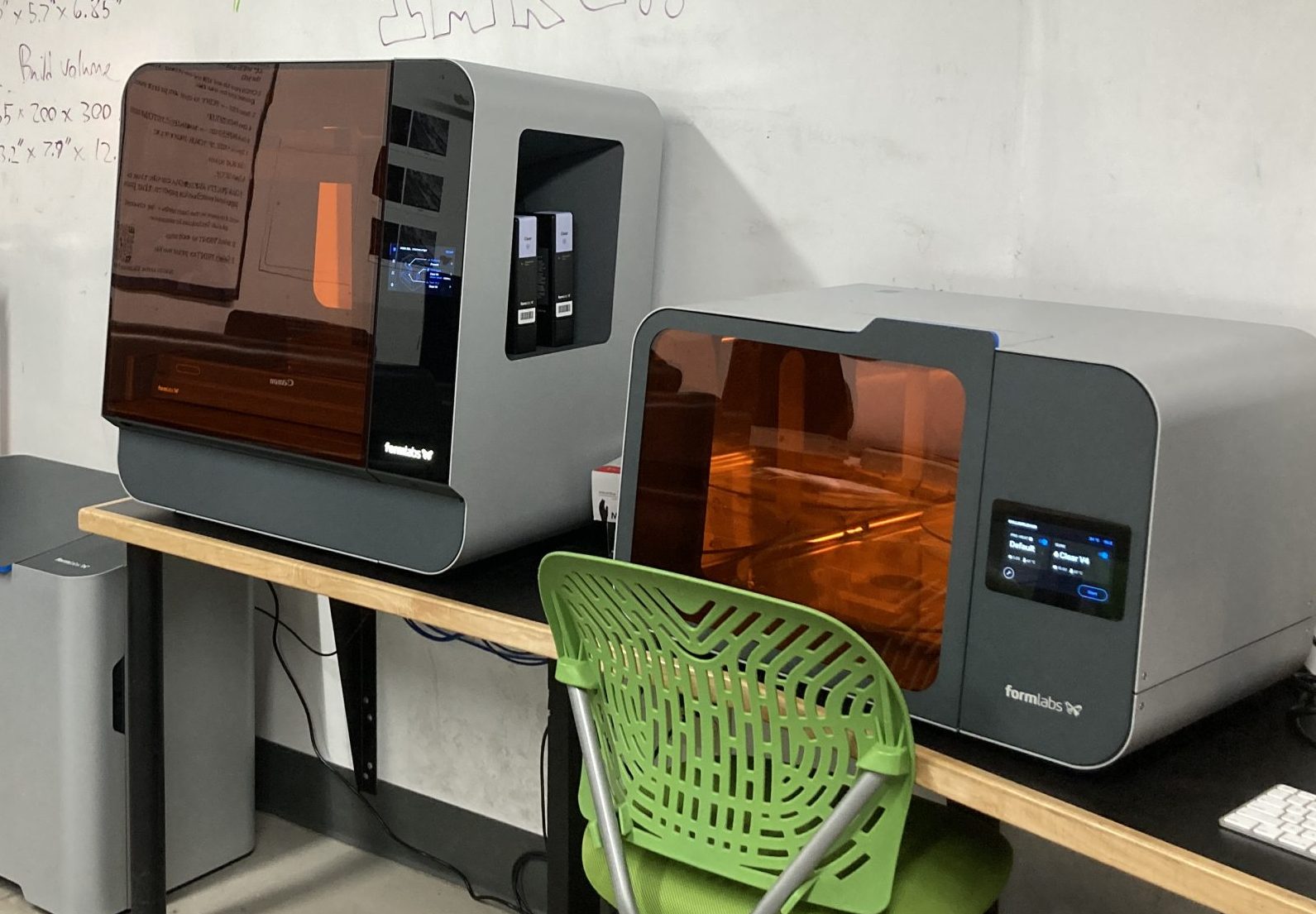 Two grey machines are on a narrow work surface next to a white wall. A green chair is in the foreground. Amber-colored glass allows the user of the machines to view items being printed and finished within them.