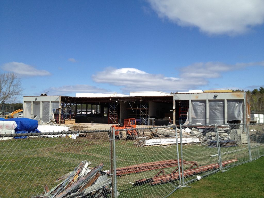 The concrete and metal framing of the former East / Stewart Commons are seen on a sunny day with clouds in the sky. Chain link fencing blocks off the construction area around the building. Construction materials and detritus can be seen in large piles. The large front windows of the building have been temporarily covered in plastic sheeting.