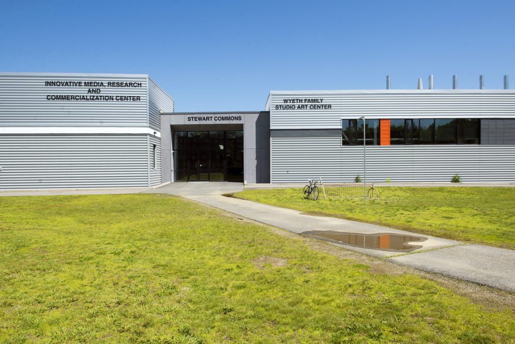 A photo of the Stewart Commons entrance as seen from the "quad. A puddle is visible on the diagonal pathed pathway to the building, but the sky is bright blue and cloudless. A bike rack with a single bicycle stands outside. On the metal cladding of the building are visible three names in black block letters: to the left, "Innovative Media, Research and Commercialization Center," in the center, "Stewart Commons," and to the right, "Wyeth Family Studio Art Center"