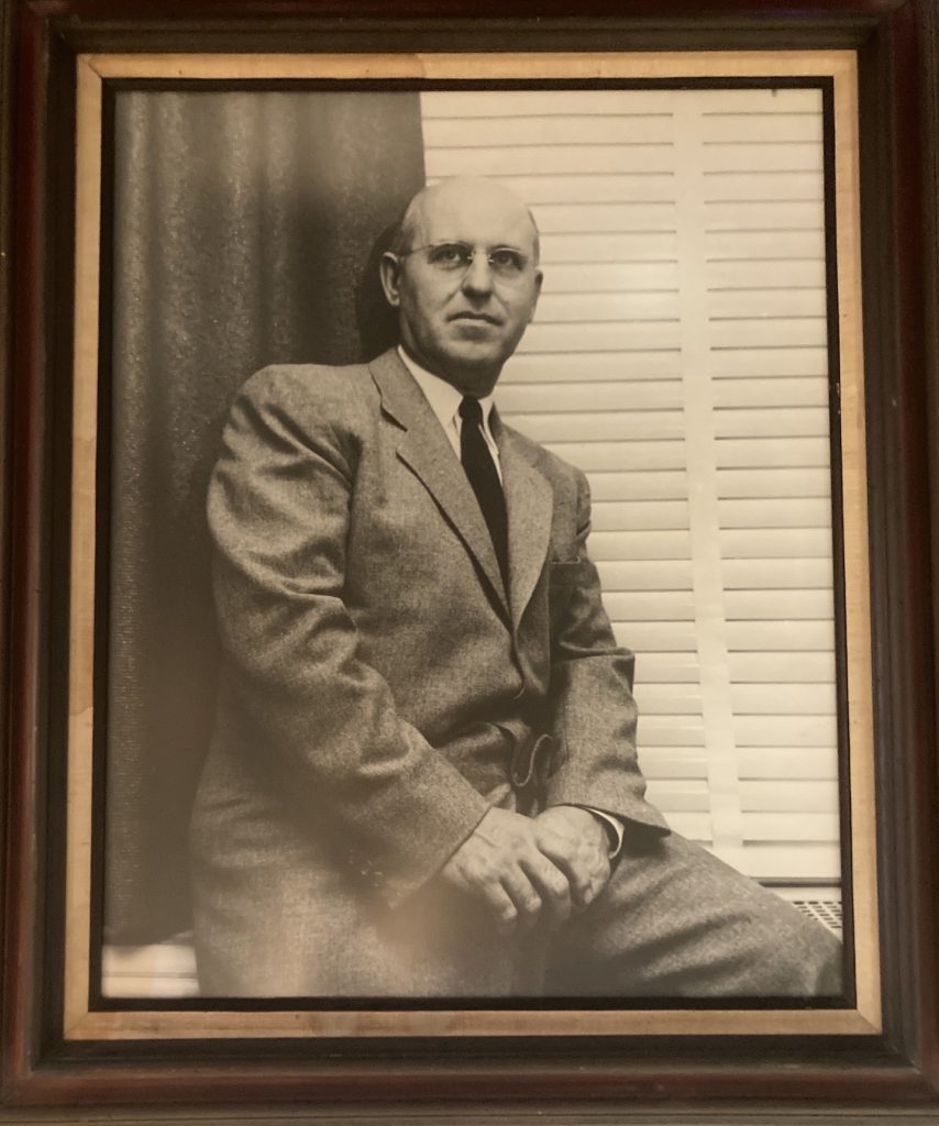 Stewart is seen sitting on a window ledge with a dark curtain and white blinds behind him. He wears a medium-toned suit and dark tie, as well as wire-rimmed glasses, and has a serious but pleasant facial expression. The black and white portrait is framed in dark wood with gold trim.