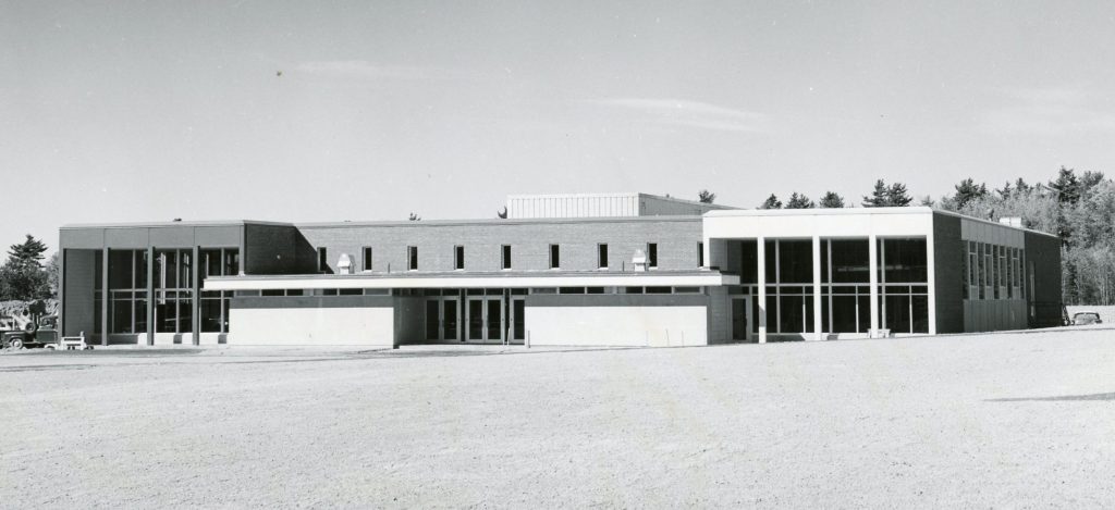 A greyscale photo of Stewart Dining Commons as it appeared after its rededication. A large, close-cut grass lawn spreads out in front of the building. A lower roof covers the front entrance doors, while two large two-story window sections can be seen to either side. A second floor over the main entrance can be seen, with a brick facade and eight smaller windows.