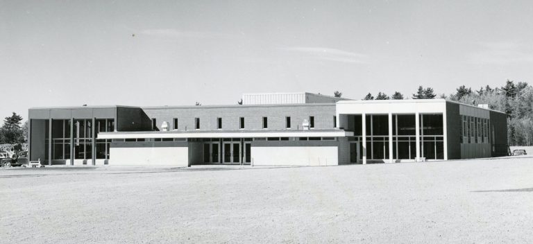 A greyscale photo of Stewart Dining Commons as it appeared after its rededication. A large, close-cut grass lawn spreads out in front of the building. A lower roof covers the front entrance doors, while two large two-story window sections can be seen to either side. A second floor over the main entrance can be seen, with a brick facade and eight smaller windows.