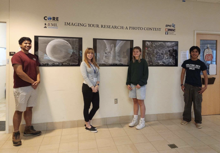 Four people stand next to a beige wall between two office doors. Staggered between them can be seen three black and white framed prints of microscopic views of different material.