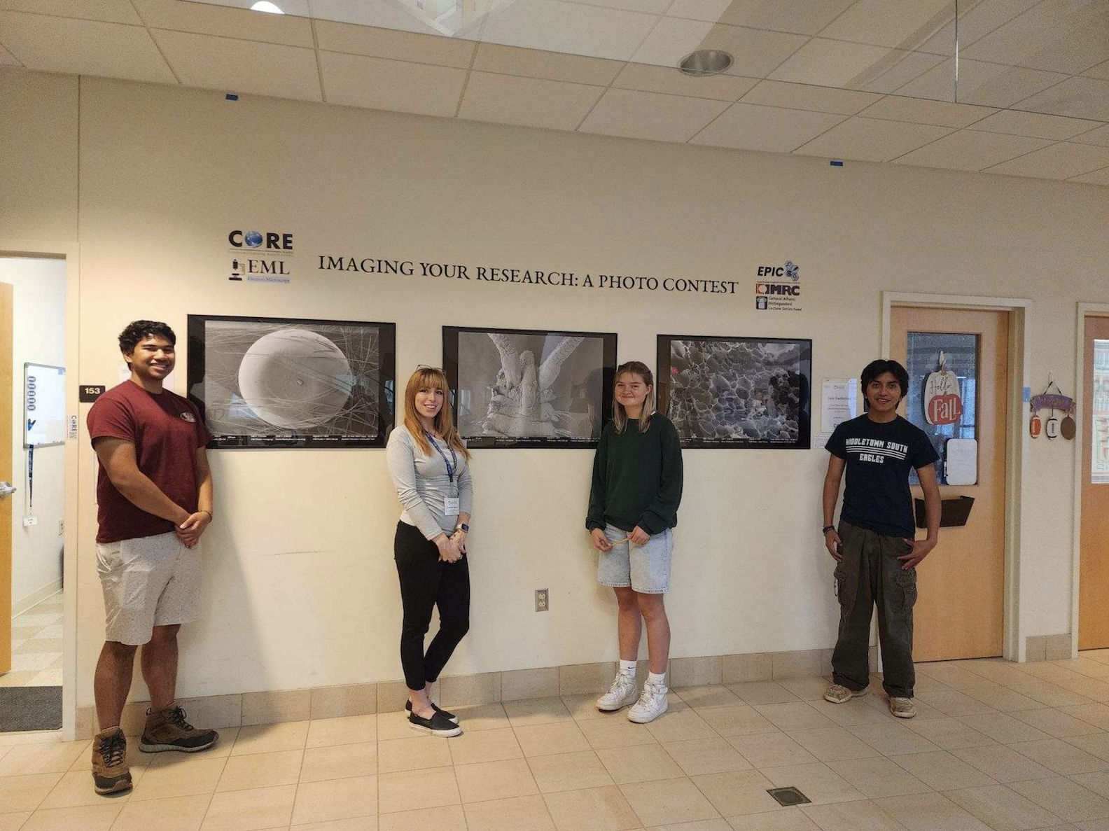 Four people stand next to a beige wall between two office doors. Staggered between them can be seen three black and white framed prints of microscopic views of different material.