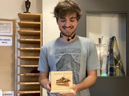 Student Bryce Butterfield looks down at an engraved wooden box project in his hands. The box shows an image of a boat, with the words "Balmy Days II" visible above it.