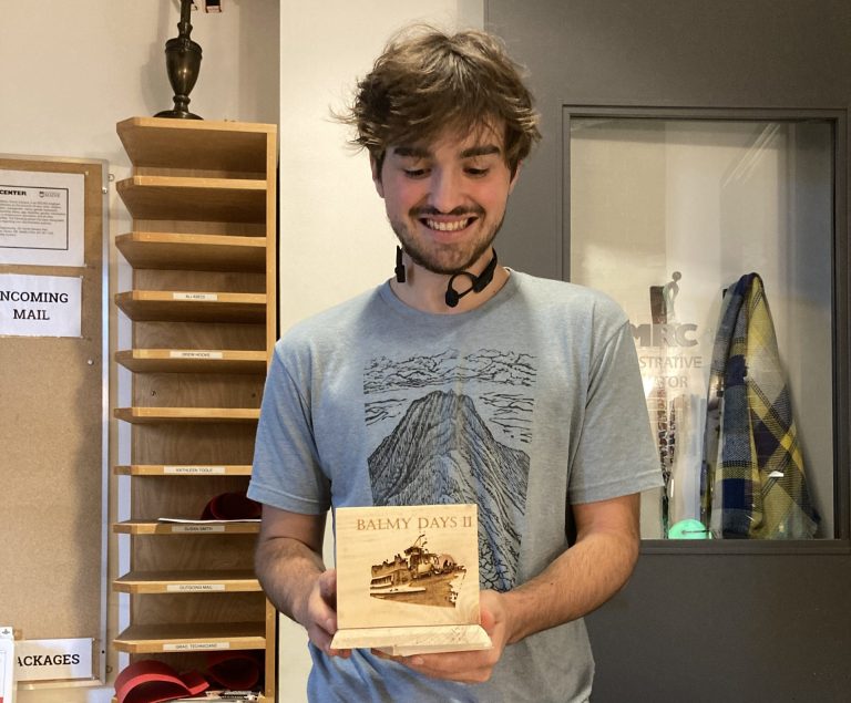 Student Bryce Butterfield looks down at an engraved wooden box project in his hands. The box shows an image of a boat, with the words "Balmy Days II" visible above it.