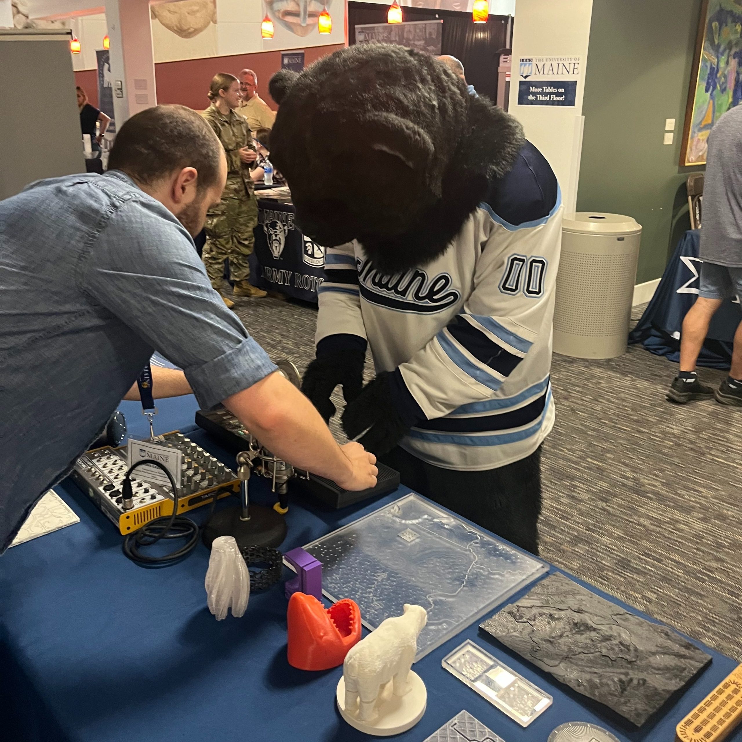 A person in a black bear mascot costume leans toward a table with a blue tablecloth. Another person demonstrates how a piece of equipment on the table works.